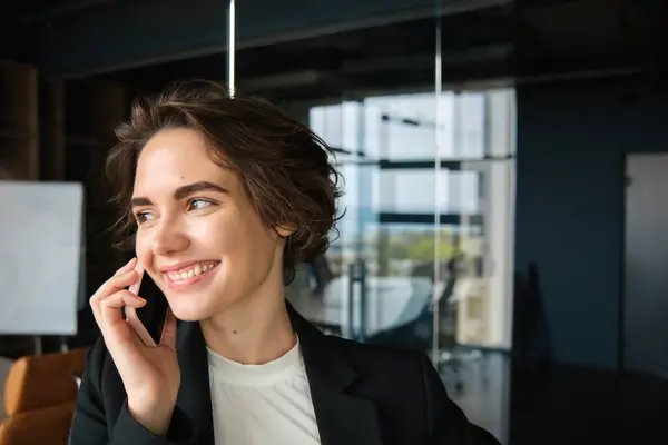 Corporate woman making phone calls, answering mobile and smiling, talking to client, having a conversation, standing in office in business suit.