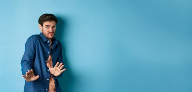 Çekingen genç adam uzak durmak istiyor, ellerini savunmaya uzatıp geri çekiliyor, iğrenç bir şeyi reddediyor, mavi arka planda duruyor..