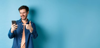 Mobil ekranı okuyan, telefonu işaret eden, online tanıtım ya da akıllı telefon uygulamasını kontrol eden, mavi arka planda duran yakışıklı bir adam resmi.