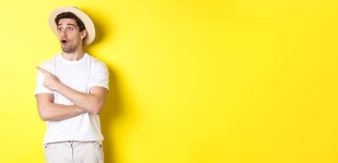 Turizm ve yaşam tarzı kavramı. Hasır şapkalı heyecanlı yakışıklı adam reklama bakıyor, sol üst köşedeki logoya bakıyor, sarı arka plan..