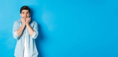 Korkmuş yetişkin bir adam korkunç bir şeye bakıyor, tırnaklarını yiyor ve korkudan titriyor, mavi arka planda duruyor..