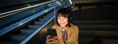 Teknoloji. Asyalı kızın portresi yürüyen merdivenin yanında oturuyor, kulaklıkla müzik dinliyor, sosyal medya kullanıyor, okuyor, umumi bir yerde video izliyor..