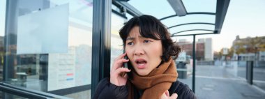 Young woman looks frustrated and disappointed, talks on mobile phone, stands on bus stop, receives bad news. Copy space