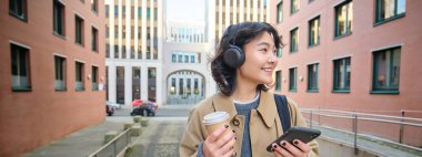 Young woman, tourist in headphones looks at smartphone, drinks coffee to go, checks mobile phone app, listens music and travels. Copy space