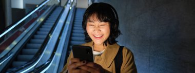 Laughing cute Korean girl on escalator, going down to tube, underground or metro, using mobile phone, watching smth funny on smartphone app.