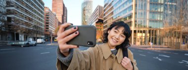 Şehir merkezindeki binanın önünde selfie çeken Asyalı genç bir kadının portresi, turistler etrafı gezerken fotoğraf çekiyor, akıllı telefon kameralarına gülümsüyor..