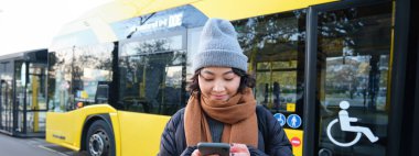 Toplu taşıma için bekleyen bir kız öğrenci resmi, akıllı telefon uygulamasında kontrol programı, belediye otobüsünün yanında duruyor. Kentsel yaşam tarzı konsepti