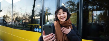 Portrait of cheerful asian girl talks on mobile phone, video chats, looks amazed at smartphone camera, stands on bus stop.