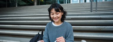 Smiling girl, graphic designer, using digital tablet and pen tool to draw, does home assignment for university, sits on stairs.