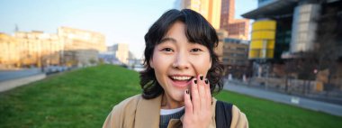 Image of korean girl with happy face, walks around town with student tablet, stands on street and smiles. Copy space