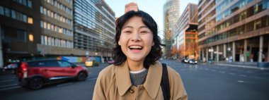 Portrait of asian girl student, stands in city centre with cars on busy street, holds digital tablet and smiles at camera.