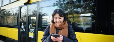 Beautiful korean girl, student on bus stop, looking at her smartphone, checking timetable, reading text message, wearing winter clothes.