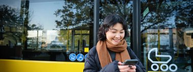 Young beautiful woman standing on bus stop, texting message on smartphone, holding mobile phone, checking her schedule, buying ticket online, wearing winter clothes.