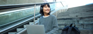 Portrait of beautiful young korean woman, student sits on stairs in public place, listens music in headphones, works on project on laptop, works remotely.