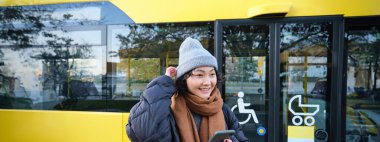 Portrait of girl standing near bus on a stop, waiting for her public transport, schecks schedule on smartphone application, holds mobile phone, wears warm clothes.