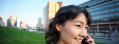 Headshot of young korean woman walks on street, talks on mobile phone, has telephone conversation, stands near campus building.