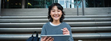 Portrait of asian girl student, hipster sitting on stairs with digital tablet and cup of coffee, draws digital art, makes design project.