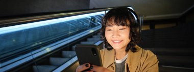 Close up portrait of smiling asian girl student, listens music in headphones and looks at mobile phone, uses smartphone, sits on staircase in public place.