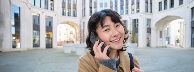 Portrait of smiling asian girl talks on mobile phone, speaks with friend on smartphone, walks in city centre, stands on street and laughs.