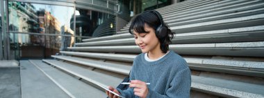 Young smiling digital artists works on her project using tablet and graphic pen, listens music in headphones and sits on stairs in public space, works on project.