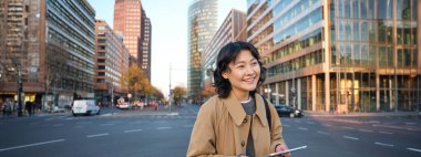 Portrait of smiling korean girl stands on busy street of city centre, holds digital tablet, casual relaxed face expression, walking around town.