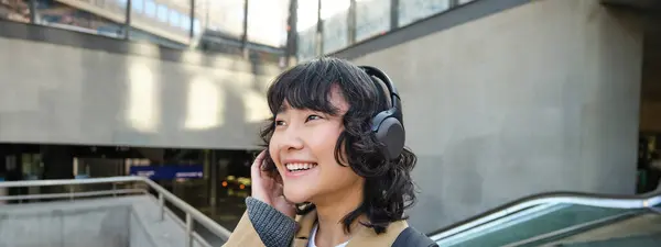 Close up portrait of Korean girl in headphones, student listens music in headphones and smiles, walks around city, tourist travelling.