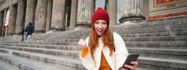 Yes, I did it. Satisfied redhead girl, sits on stairs on street and looks pleased, celebrates achievement, makes fist pump, holds mobile phone.