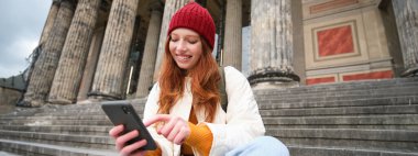 Stylish young redhead woman, talking on mobile phone app, using social media application, looking for something online on smartphone, sits on stairs outdoors.