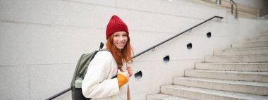 Happy young girl, redhead tourist in red hat with backpack, walks around town, explores city, backpacking around europe, travelling alone.