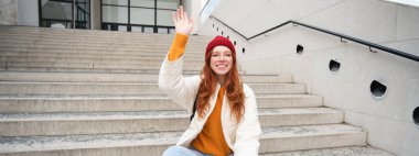 Smiling young redhead girl waits someone on stairs outdoors, waves hand at friend, holds smartphone, says hello. People and lifestyle concept