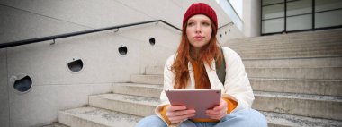 Young stylish girl, redhead female students sits on stairs outdoors with digital tablet, reads, uses social media app on gadget, plays games while waits on street.