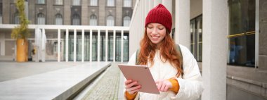 Happy redhead girl in red hat, walks around city with digital tablet, connects to public internet wifi and looks for route, looks at map on her gadget.