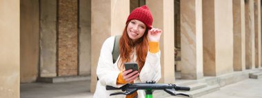 Beautiful young woman, tourist with backpack, rents e-scooter to travel around town, uses mobile phone application.