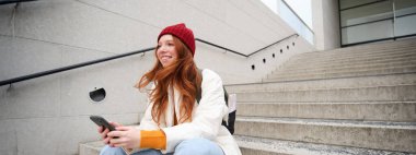 Mobile phones and people. Young stylish redhead girl sits on stairs with telephone, uses smartphone app, reads smth online.