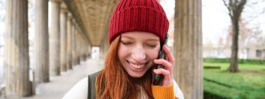 Portrait of redhead european girl in red hat, makes a phone call, walks in city and talks to friend on smartphone. Copy space