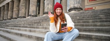 Redhead female tourist talks on mobile phone, video chatting with friends, says hi and waves hand at smartphone camera.