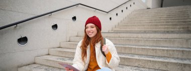 Happy redhead woman in red hat, sits on stairs outdoors, uses tablet, connects to wifi near public building, reads digital book while waits on street.
