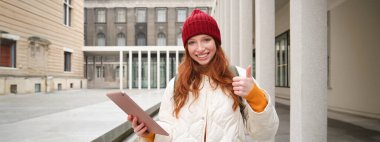 Stylish ginger girl, tourist walks with digital tablet around city, woman connects to iternet on her gadget, looking up information, texting message.