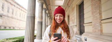 Kızıl saçlı, gülümseyen bir kadın, gezerken cep telefonu uygulaması kullanan bir turist, ziyaret edecek bir yer arıyor, akıllı telefon uygulaması ile gezmeyi keşfediyor..