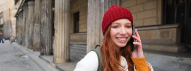 Smiling redhead female tourist talks on mobile phone and walks around city. Happy student in red hat calls friend, stands on street and uses smartphone.