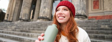 Portrait of redhead woman drinking from thermos, sitting on street stairs and enjoys hot drink from flask.