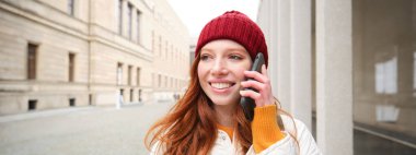 Happy redhead woman, girl with phone talks, has conversation on mobile app, uses internet to call abroad with smartphone app, laughing and smiling.