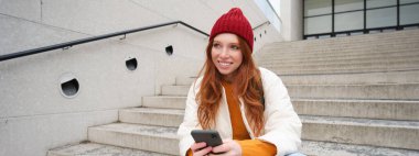 Redhead girl in red hat, sits on stairs and uses mobile phone. Modern woman holding smartphone, texting message, using telephone application outdoors.