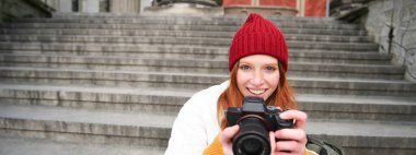 Young student, photographer sits on street stairs and checks her shots on professional camera, taking photos outdoors.