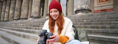 Young student, photographer sits on street stairs and checks her shots on professional camera, taking photos outdoors.