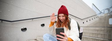 Happy college girl talks with friends on video chat smartphone app, sits on stairs outdoors uses her mobile phone application, waves hand at telephone camera.