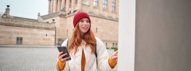Smiling redhead girl tourist, walks around city and explores popular landmarks, sightseeing, holding smartphone, looking at her mobile app and checks with city map.
