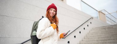 Redhead girl, young woman tourist with backpack, holds smartphone, looks for route on mobile application, searches for hotel on phone map, smiles happily.