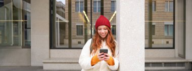 Stylish urban girl using mobile phone app, standing in city, waiting for taxi, looking at smartphone application, texting message.