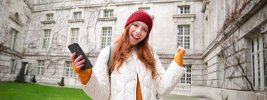 Cheerful redhead girl winning, celebrating victory, read great news on mobile phone and jumping from joy, saying yes, standing outside building in yard.